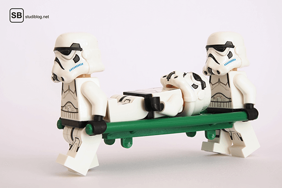 2 Lego-Sturmtruppler von Stars Wars tragen einen weiteren Sturmtruppler mit einer grünen Liege nach einem Unfall - Die Berufsunfähigkeitsversicherung.